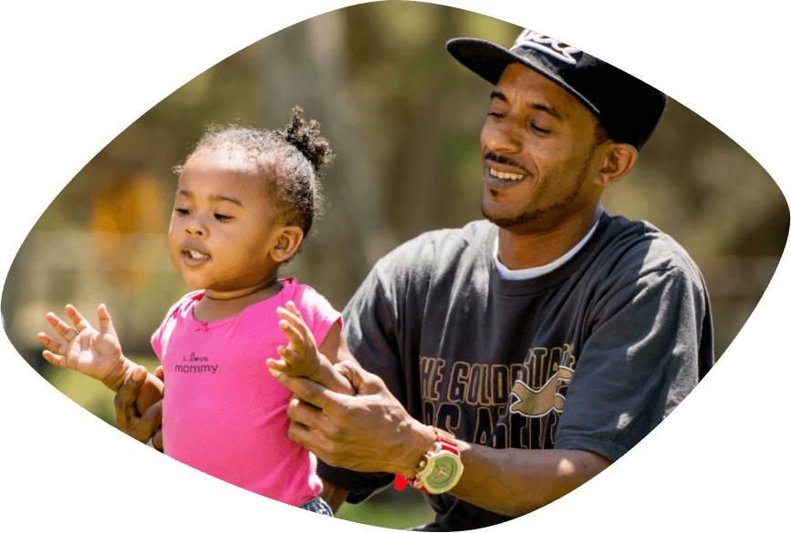 Un sonriente papá afroamericano, vestido en camiseta y gorra de béisbol, le está ayudando a su sonriente hijita a pararse.