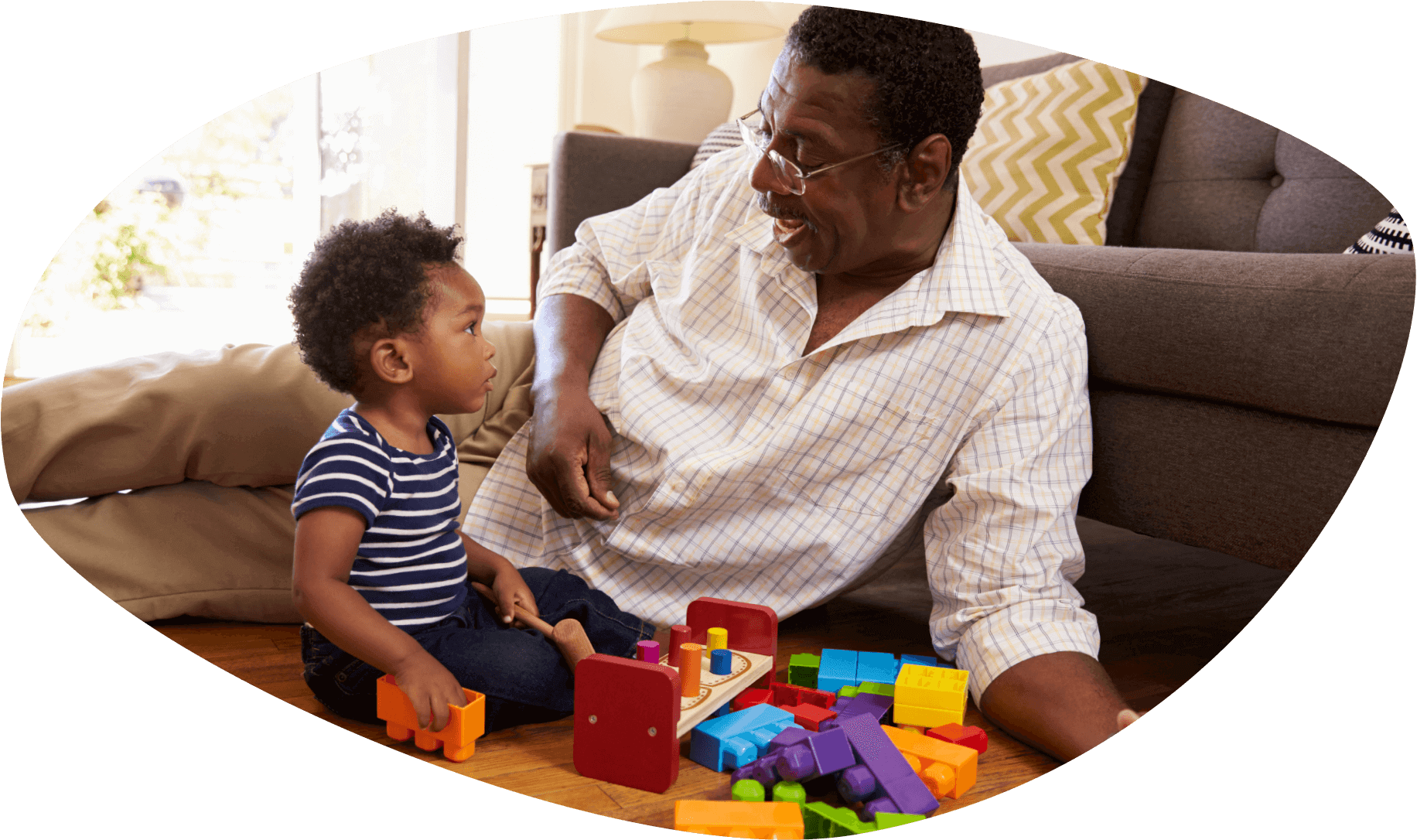 Un niñito afroamericano está viendo cómo se ríe su papá mientras que están sentados en el suelo jugando con unos legos gigantes.